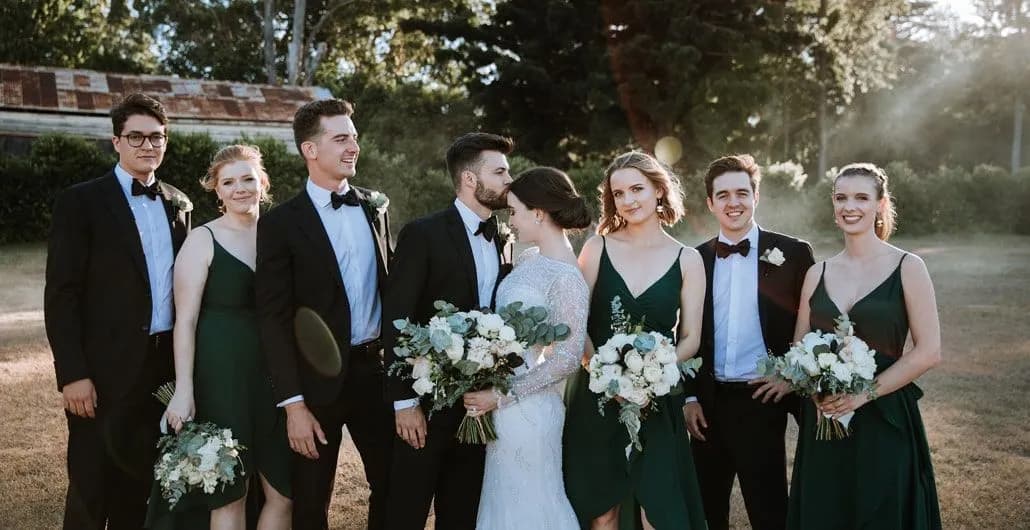 A bride in a white dress and groom in a black tuxedo stand in the center, surrounded by three bridesmaids in green dresses holding bouquets and three groomsmen in black tuxedos. They appear to be outdoors with greenery and an old building in the background.