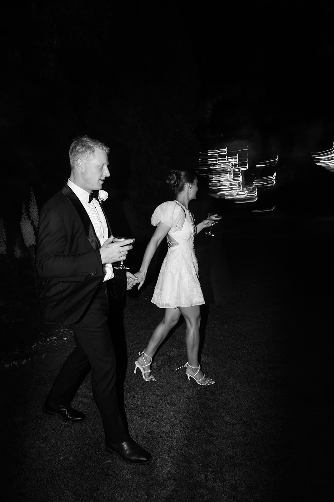 a newly married couple walking through the grounds of gabbinbar at night holding drinks