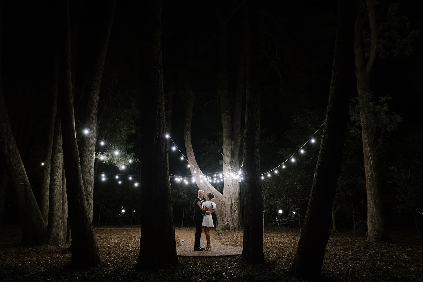 A couple embraces under a canopy of string lights hanging among tall trees in a dimly lit forest at night. The lights create a romantic, intimate atmosphere as they stand closely together on a path surrounded by darkness.