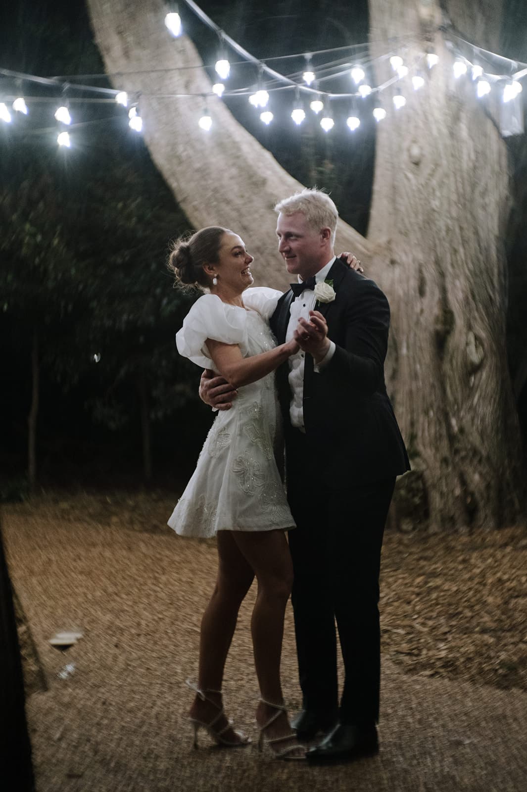 A couple dressed formally, with the woman in a short white dress and the man in a black suit, dance happily under string lights at night in an outdoor setting surrounded by trees.