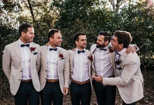A group of five men dressed in beige suits with white shirts, and blue bow ties stand together outdoors. They all have dark trousers and red boutonnières. They are smiling, with their arms around each other, appearing relaxed and cheerful amidst the greenery.