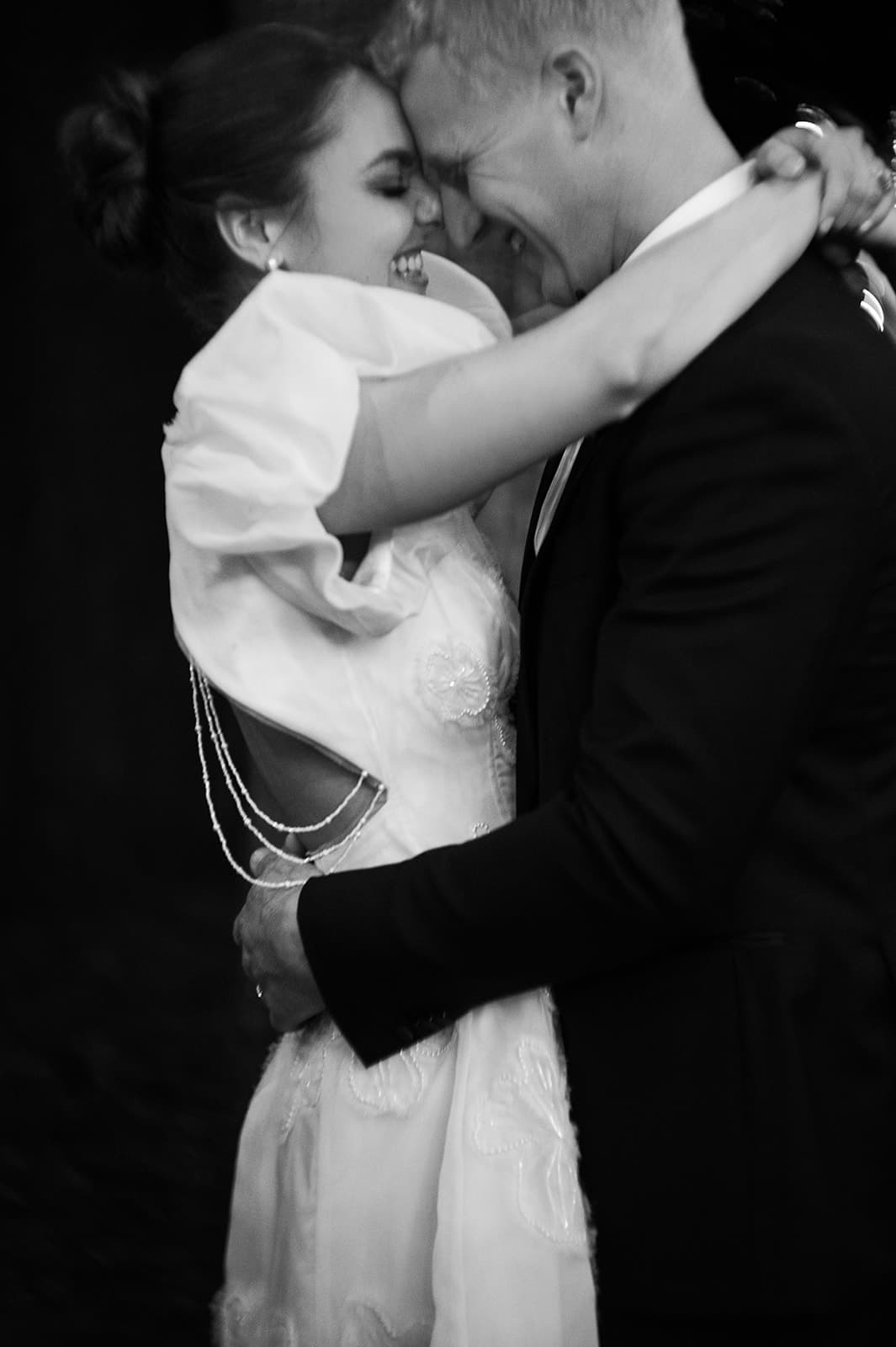 A bride and groom are joyfully embracing in a close-up black and white photo. The bride's elegant dress has delicate beadwork and a shoulder detail, while the groom wears a dark suit. They are smiling and appear caught in a moment of happiness and closeness.