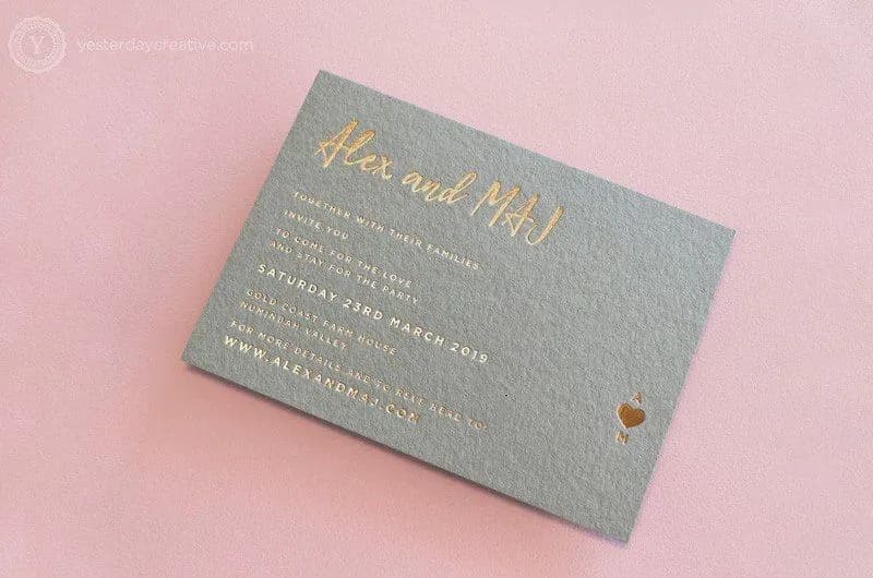 A wedding invitation card on a pink background. The card has metallic gold text with the names "Alex and Mai" in cursive at the top. Below, event details are included, such as date, venue, and a website. A small heart icon is printed in the bottom right corner.