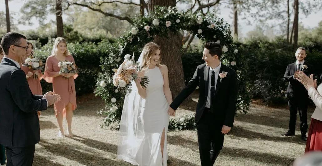 A bride in a white dress and a groom in a black suit walk hand in hand, smiling, under a floral arch in an outdoor setting. Bridesmaids in pink dresses and groomsmen in suits stand nearby, clapping. Trees and greenery surround the happy scene.