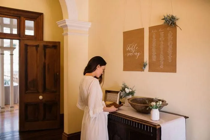 A woman in a white dress stands in a warmly lit hallway, looking at a book on a wooden table. Above the table, two large pieces of brown paper with calligraphy and floral decorations are hanging on the wall. A doorway and a window are visible in the background.
