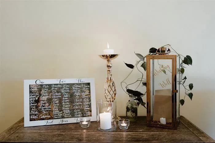 A table displays a wedding seating chart, candles, and a lantern. The seating chart lists guests' names written in white on a glass pane. Several lit candles, including one in a glass holder, add a warm glow to the arrangement. A leafy plant is in the background.