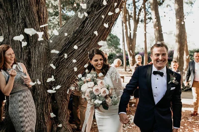 A couple, dressed in formal wedding attire, walks under a large tree as rose petals are showered upon them. The bride holds a bouquet of roses and the groom smiles broadly. Guests in the background look on, smiling and celebrating.