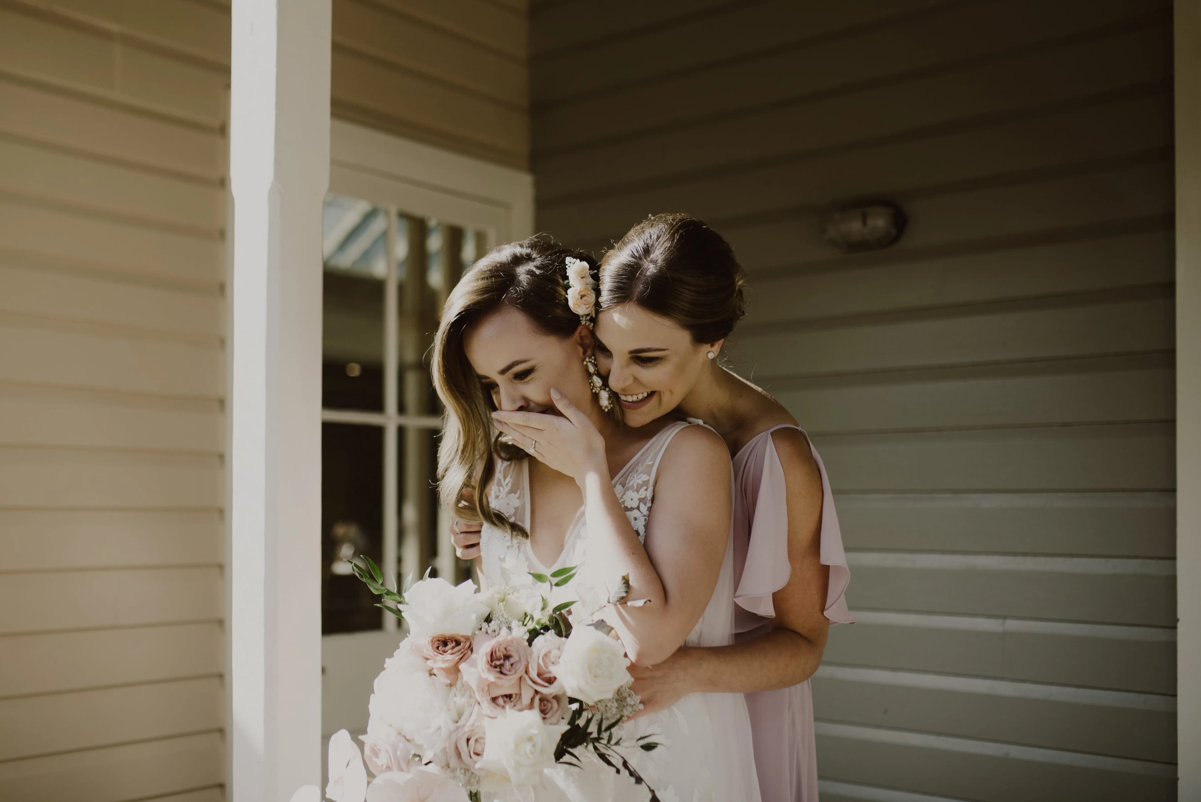 Two women, one in a white wedding dress holding a bouquet of flowers and the other in a light purple dress, are standing on a porch. They are smiling and laughing together, with the woman in the purple dress hugging the bride from behind.