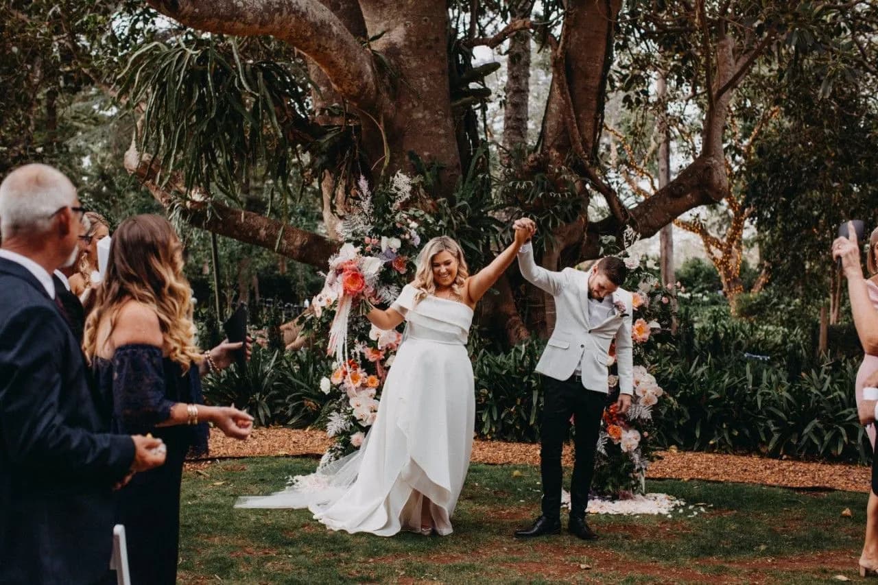 A bride and groom celebrate their wedding ceremony outdoors, holding hands and raising their arms in joy. They stand beneath a large tree adorned with flowers, surrounded by happy guests. The bride wears a white dress, and the groom wears a light-colored suit.