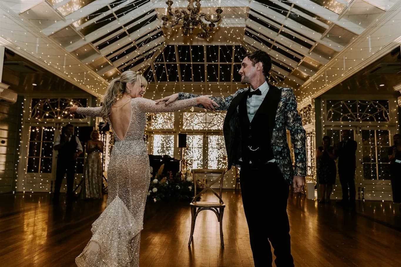 A couple dances under twinkling fairy lights in a beautifully decorated venue. The woman wears a sparkly, backless gown, and the man wears a dark suit with a patterned jacket. They hold hands, smiling at each other as they twirl. A chandelier hangs from the ceiling.