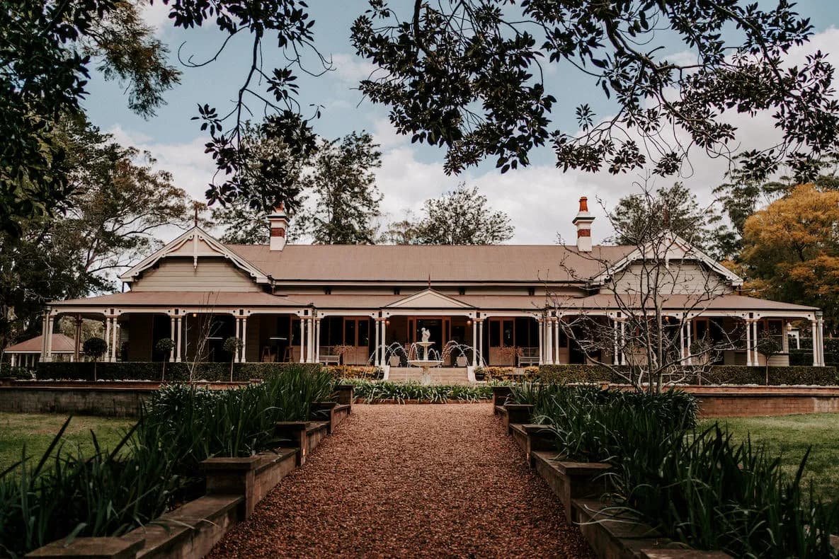 A scenic view of a large, historic-style house surrounded by a lush garden. The home has a veranda with white railings and pillars, a gabled roof with multiple chimneys, and is framed by tree branches. A gravel pathway leads to the entrance through well-manicured greenery.