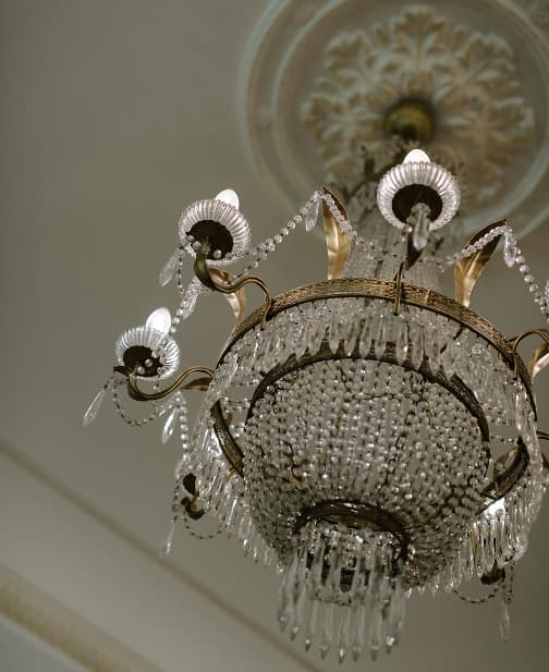 A luxurious, ornate chandelier adorned with intricate crystal droplets and gold accents hangs from a decorative ceiling with a floral rosette. The chandelier emits a soft glow, casting a sophisticated ambiance in the elegantly designed room.