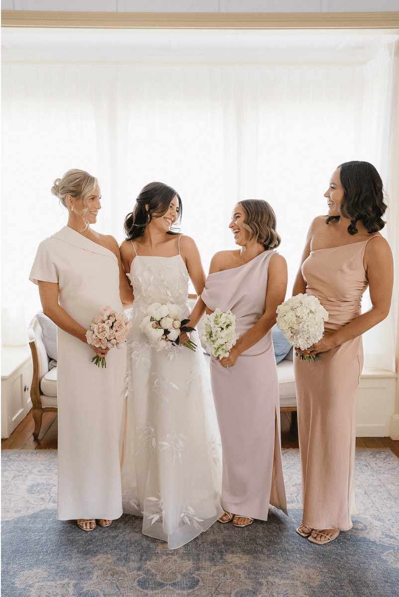 A bride in a white floral gown stands with three bridesmaids in pastel dresses. Each holds a bouquet and smiles while looking at one another. They are in a well-lit room with large windows and a patterned rug.