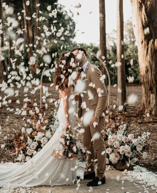 A couple kisses at an outdoor wedding ceremony. They are surrounded by a shower of flower petals. The bride wears a white gown and holds a bouquet, while the groom is in a tan suit. Behind them, there are floral arrangements and tall trees.