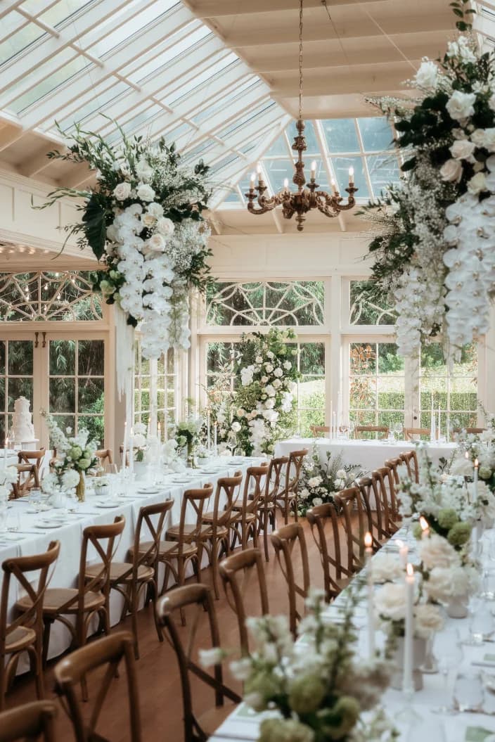 A beautifully decorated wedding reception with long tables adorned with white flowers and greenery. Elegant chandeliers hang from a glass ceiling, and large floral arrangements cascade down from above, creating a sophisticated and airy atmosphere.