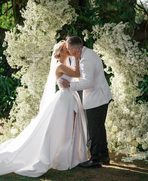 A couple shares a kiss in front of lush greenery and white floral arrangements at an outdoor wedding. The bride wears a flowing white gown, and the groom is in a white suit jacket and black pants. Sunlight filters through the foliage.
