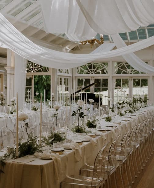 A beautifully decorated wedding reception hall with elegant white drapery hanging from the ceiling. The long tables are set with white linens, tall candles, and floral centerpieces. Ghost chairs are neatly arranged around the tables.