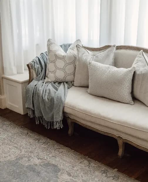 A cozy living room setting with a vintage-style white sofa adorned with multiple patterned pillows and a light blue throw blanket. White curtains give a soft, natural light ambiance to the room, complemented by a light-colored rug.