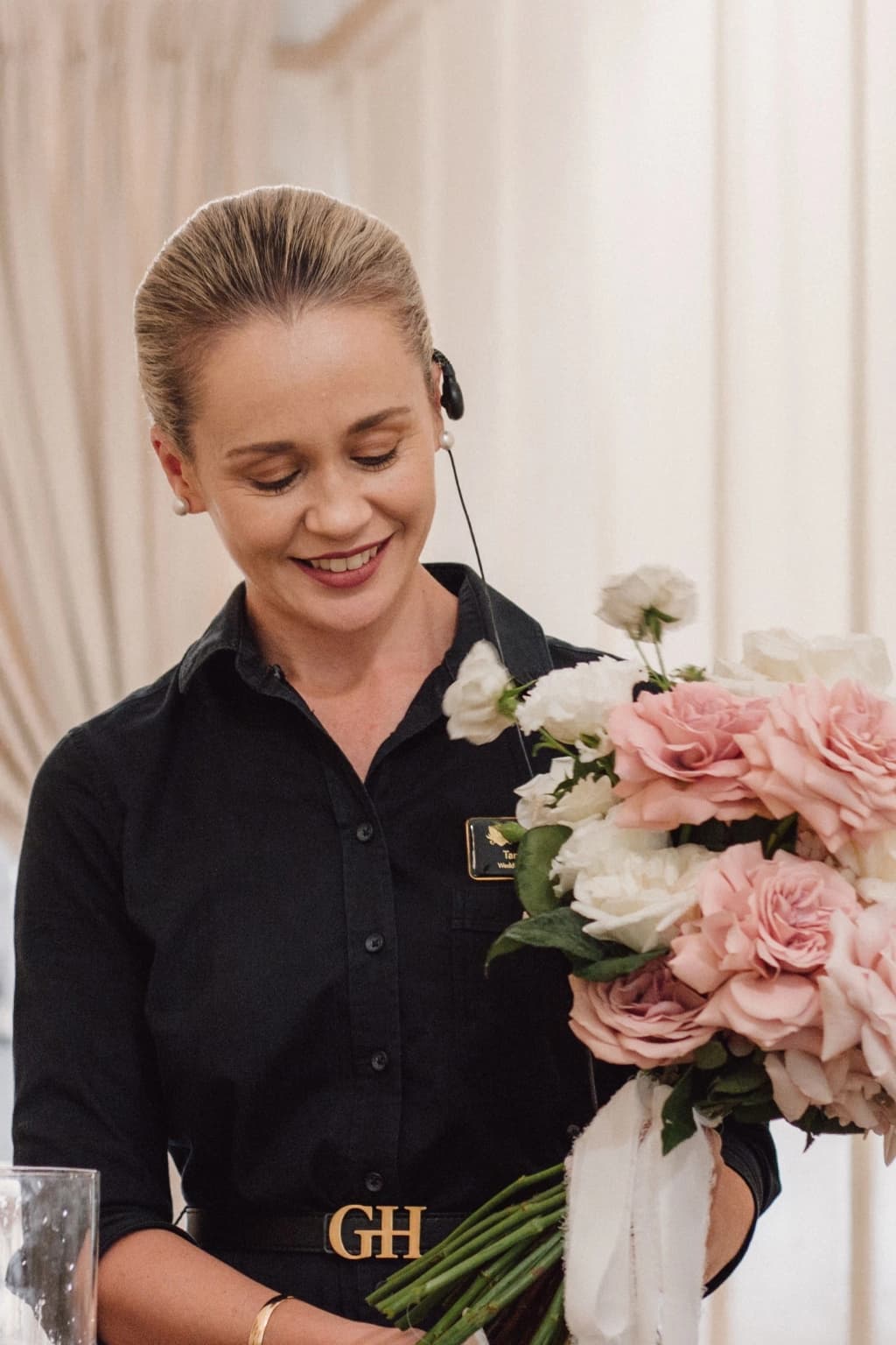 A person smiles while holding a bouquet of pink and white flowers. They are wearing a dark shirt with a headset on and are positioned in front of cream-colored curtains.