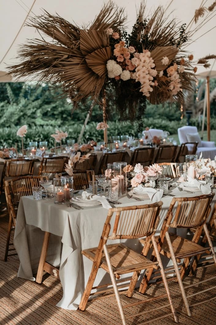 A beautifully decorated outdoor wedding reception with wooden chairs and tables adorned with white linens, candles, and floral arrangements. Above, there is a hanging decoration of dried palm leaves and pink flowers.