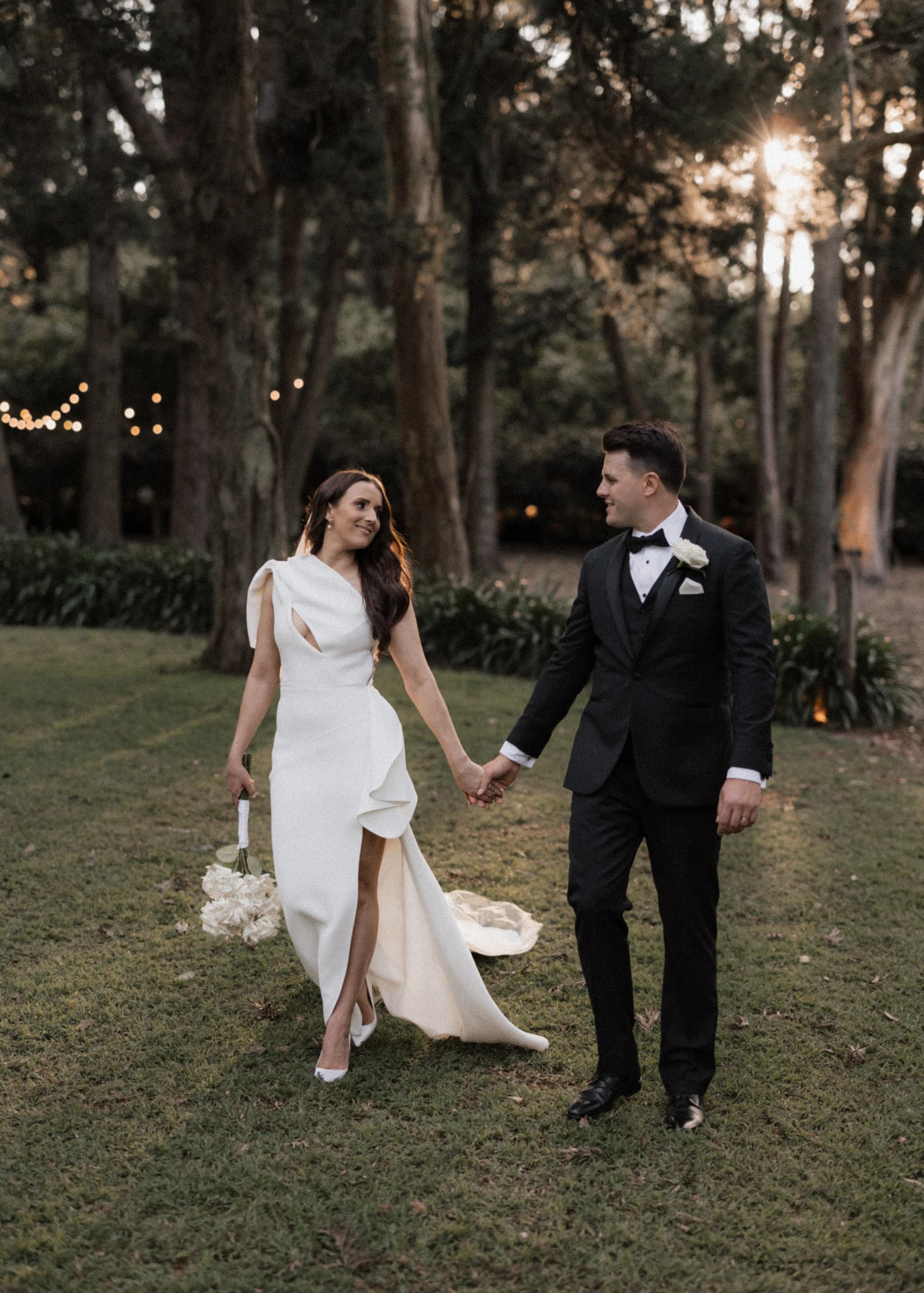 A bride and groom walk hand-in-hand on a grassy lawn during an outdoor wedding. The bride wears a white dress and holds a bouquet, while the groom is in a black suit. Trees and string lights are visible in the background.
