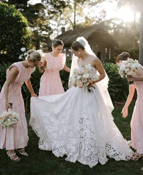 A bride in a white lace gown holds a bouquet and is surrounded by three bridesmaids in light pink dresses, who help arrange her dress. They stand on grass, with trees and a small building in the background, illuminated by soft sunlight.