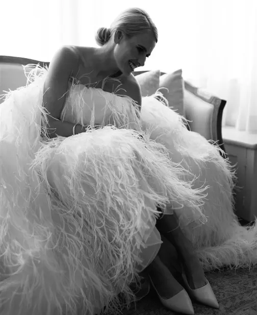 A person sitting and smiling, wearing a strapless, feathered gown. The setting appears to be indoors, next to a window with sheer curtains. The image is in black and white, creating a soft, elegant atmosphere.